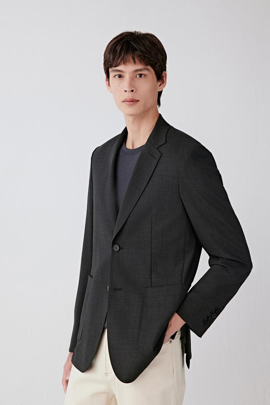 Man wearing a charcoal black twist wool jacket with a tailored straight cut and notched lapel, styled with light trousers; front three-quarter view showing the clean lines, structured shoulders and natural drape of the fabric.