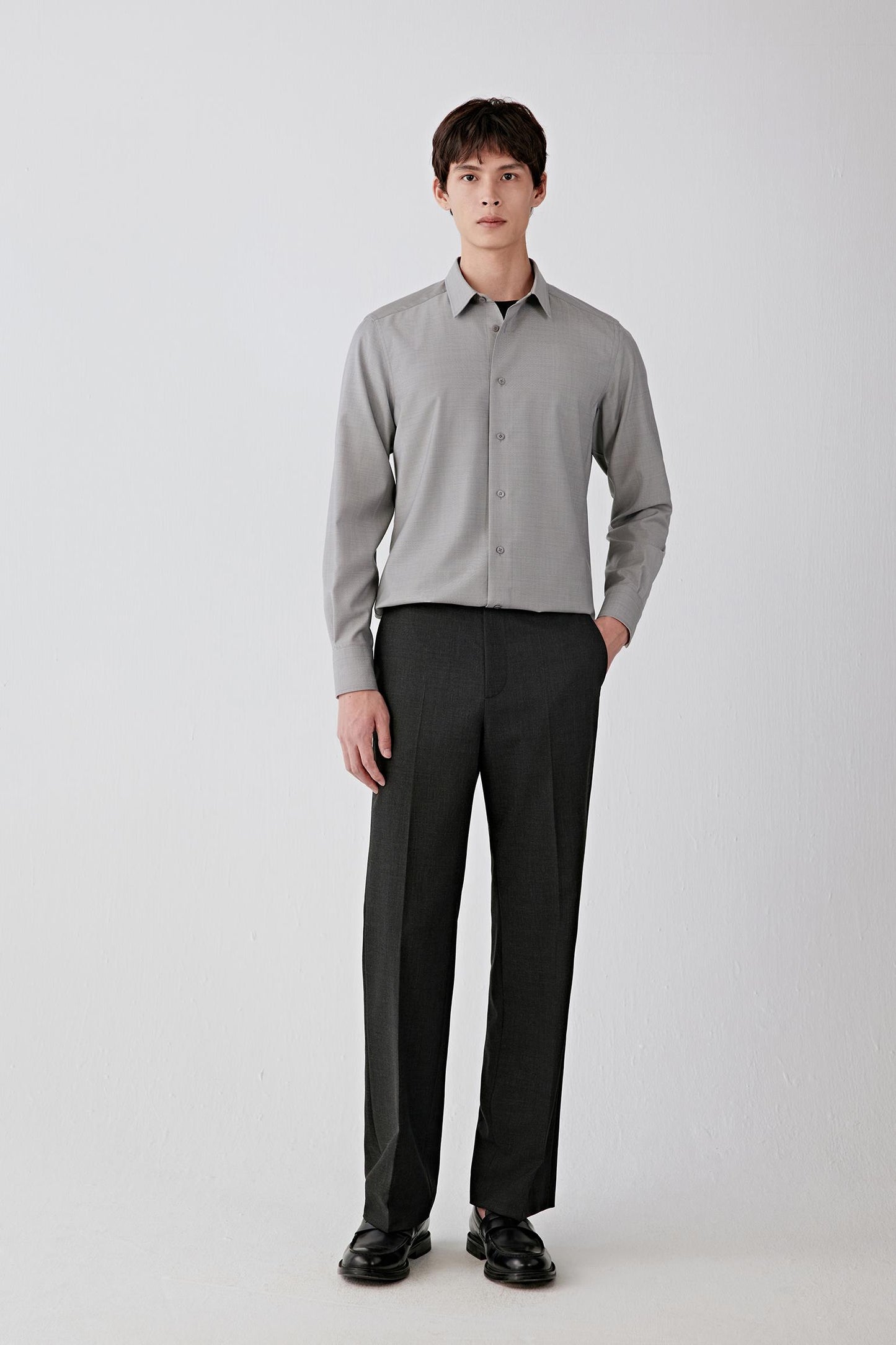 Full-length front view of a male model wearing charcoal black straight-leg trousers in twist wool, styled with a light grey knit and black shoes, showing a clean waistband and fluid drape.