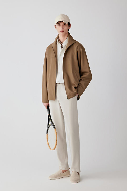 Person wearing a brown jacket, white shirt, beige pants, and cap, holding a tennis racket on a white background