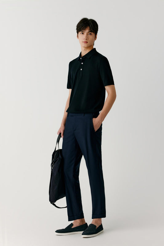 A man modeling a black short-sleeve polo shirt and navy blue trousers, holding a black duffel bag against a light gray studio background.