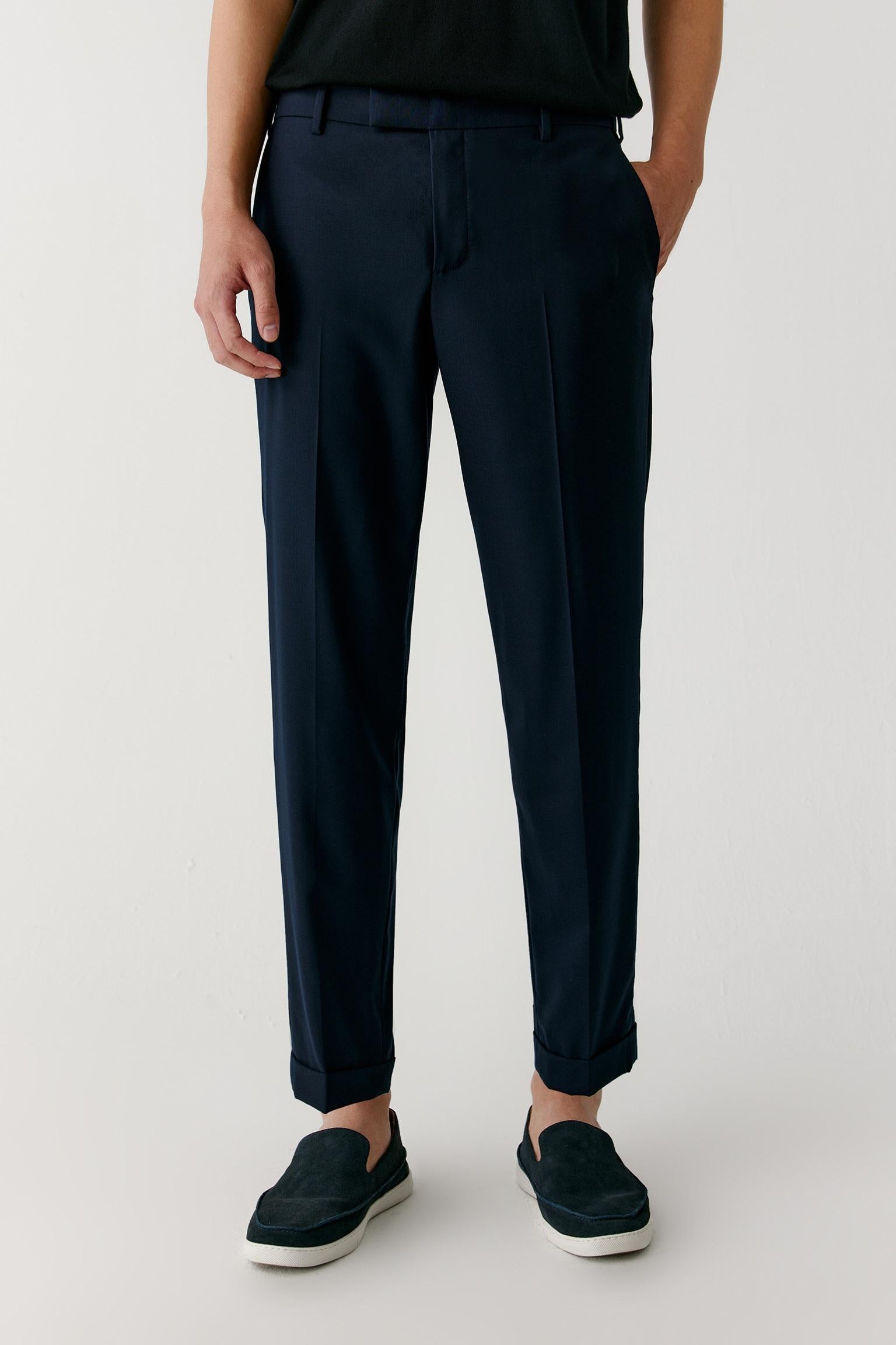 A person wearing dark navy tailored trousers with a black top and dark slip-on shoes, photographed against a clean light gray studio background.