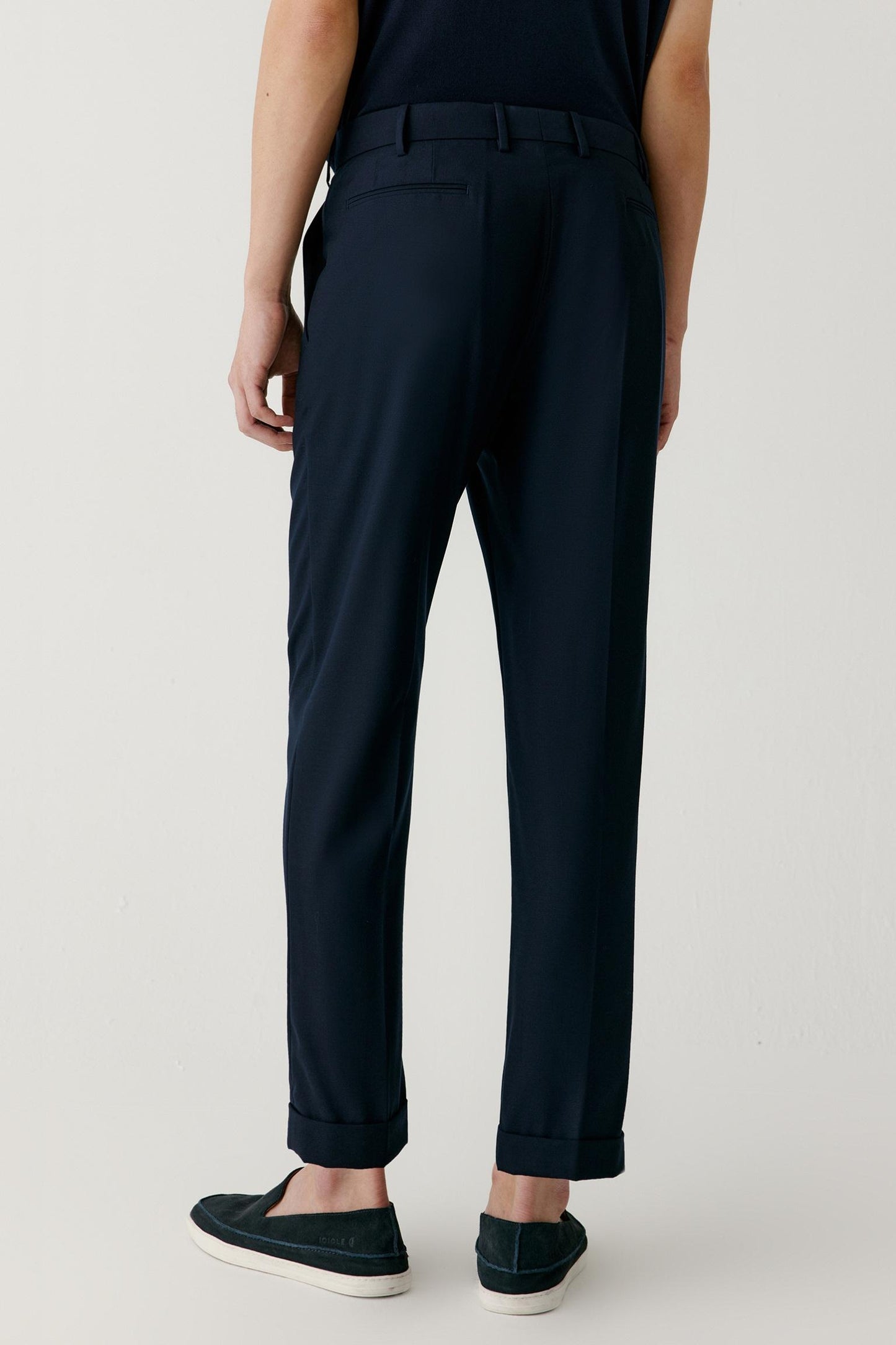 A person shown from the back wearing dark navy trousers with a belt, paired with dark slip-on shoes, against a plain light gray studio background.