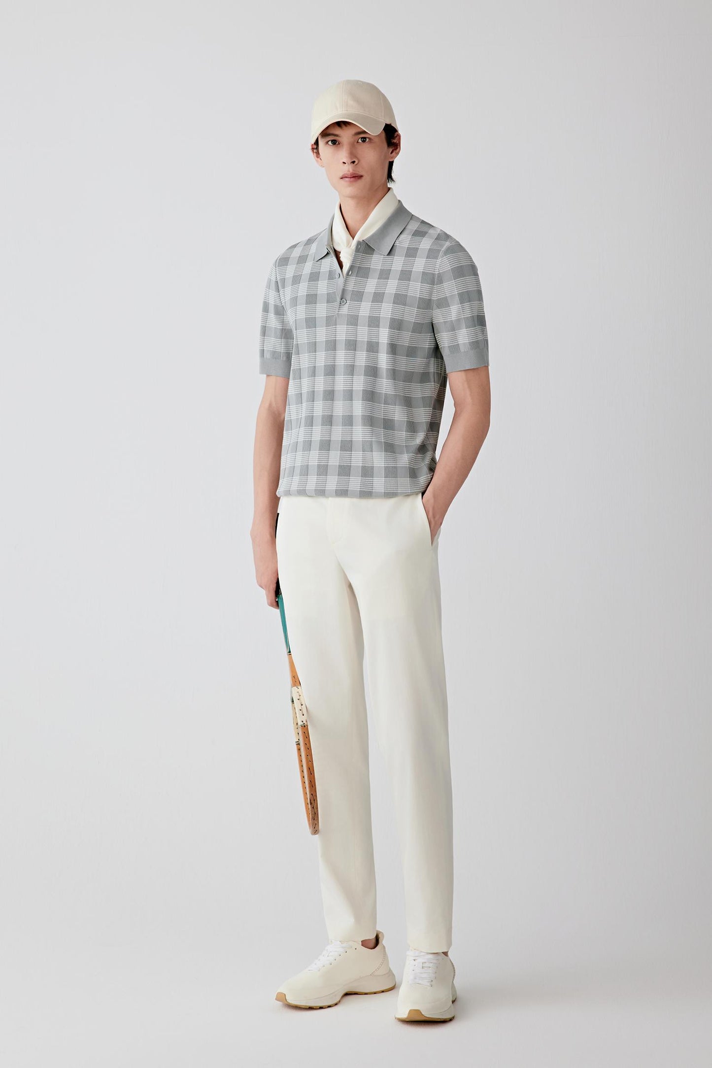 Man wearing a light grey cotton knit polo-style pullover with a subtle small-scale checked pattern, paired with straight off-white trousers, light shoes and a cap; full-length front view showing a relaxed straight cut, slightly dropped shoulders and a soft, fluid drape.