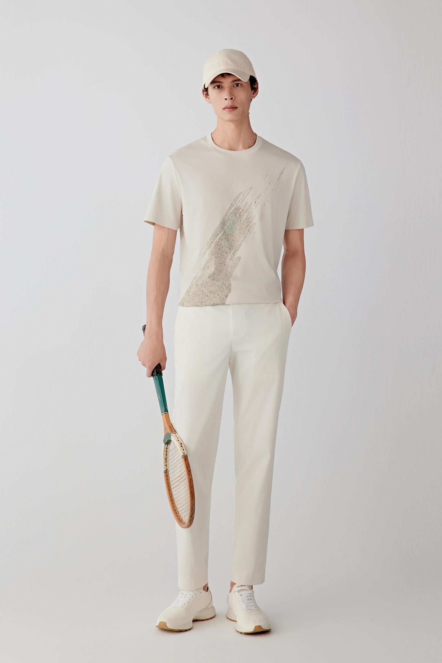 Man wearing a light nude cotton T-shirt with a straight, relaxed fit, paired with off-white trousers, light shoes and a cap; full-length front view showing the soft structure and clean silhouette.
