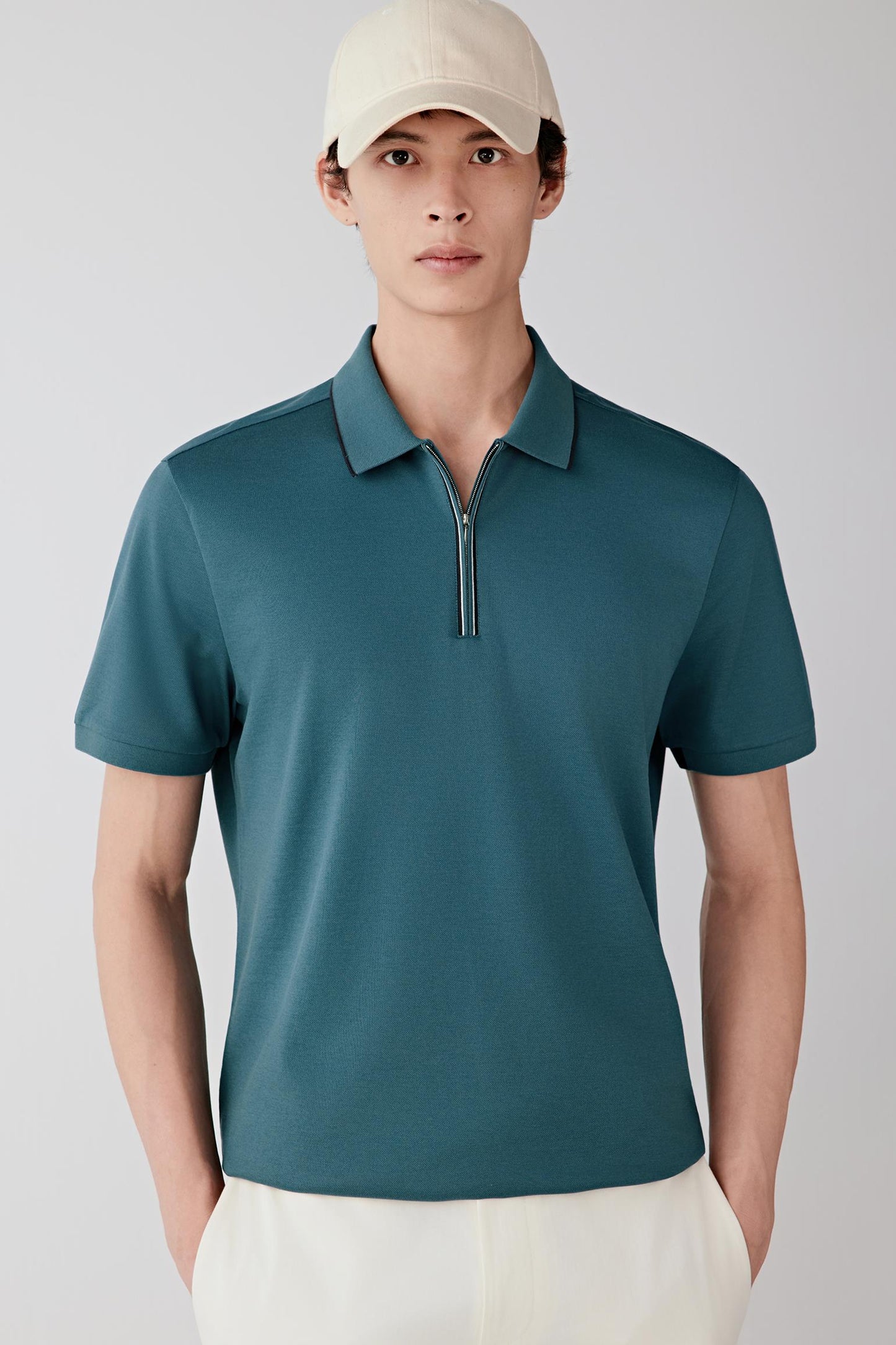 Man wearing a blue-green silk-cotton piqué polo shirt with a straight, relaxed fit, paired with off-white trousers and a light cap; full-length front view showing the structured yet breathable piqué texture and clean silhouette.