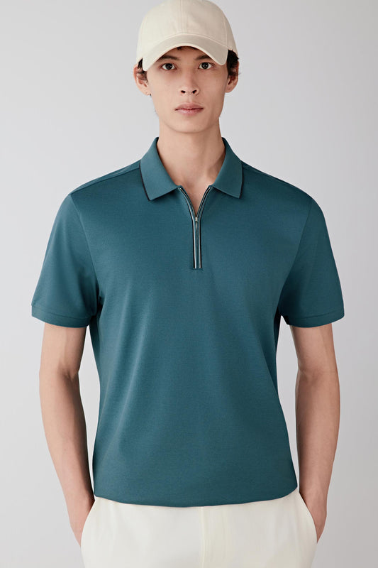Man wearing a blue-green silk-cotton piqué polo shirt with a straight, relaxed fit, paired with off-white trousers and a light cap; full-length front view showing the structured yet breathable piqué texture and clean silhouette.