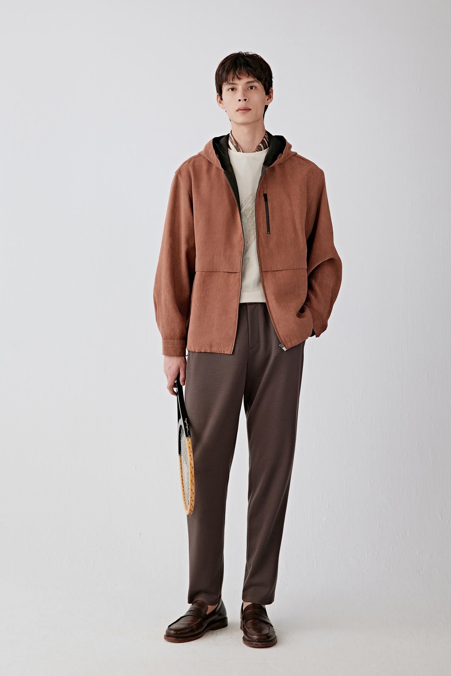 Full-length front view of a male model wearing a rust-brown jacket with hood, styled over dark trousers, showing zip fastening and relaxed cut.