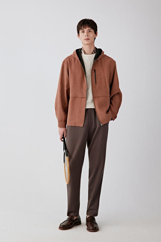 Full-length front view of a male model wearing a rust-brown jacket with hood, styled over dark trousers, showing zip fastening and relaxed cut.