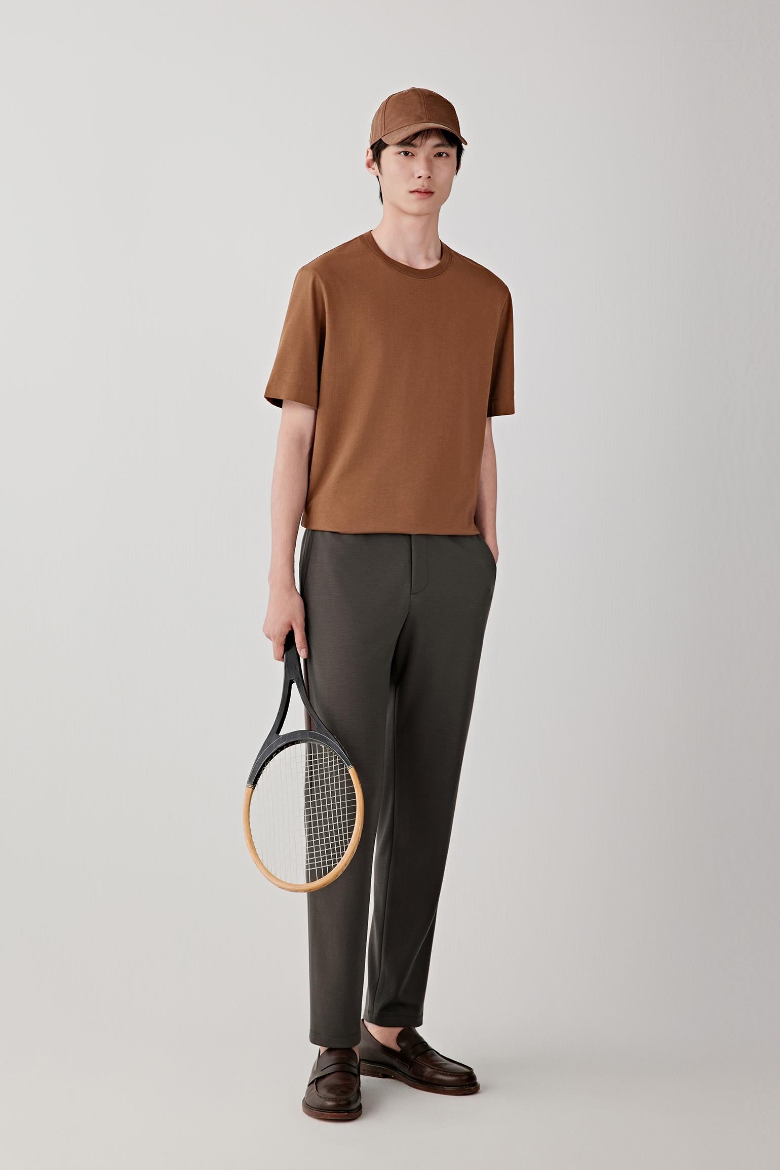 Man wearing a chestnut brown cotton jersey T-shirt with a straight, relaxed fit, paired with dark grey trousers, light shoes and a cap; full-length front view showing the clean silhouette and even drape.