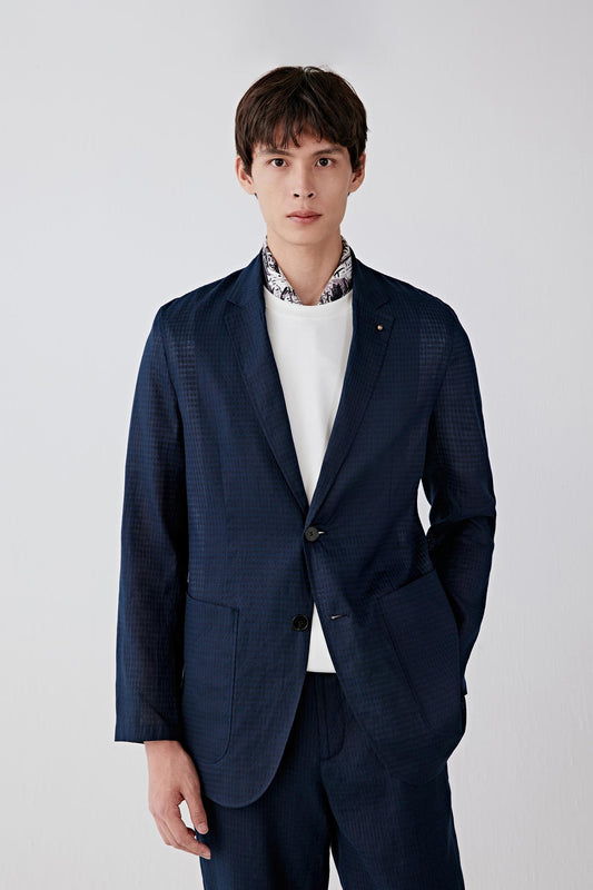 Front view of a man wearing a jacket in a navy linen and cotton blend with a subtle black check pattern, highlighting the lightweight texture, soft structure and relaxed tailoring of the fabric.