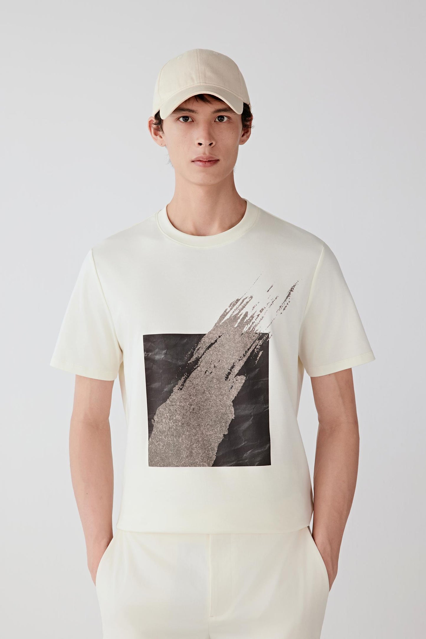 Three-quarter view showing the T-shirt paired with light trousers. The image emphasizes the balanced proportions, short sleeves and structured drape created by the compact interlock knit.
