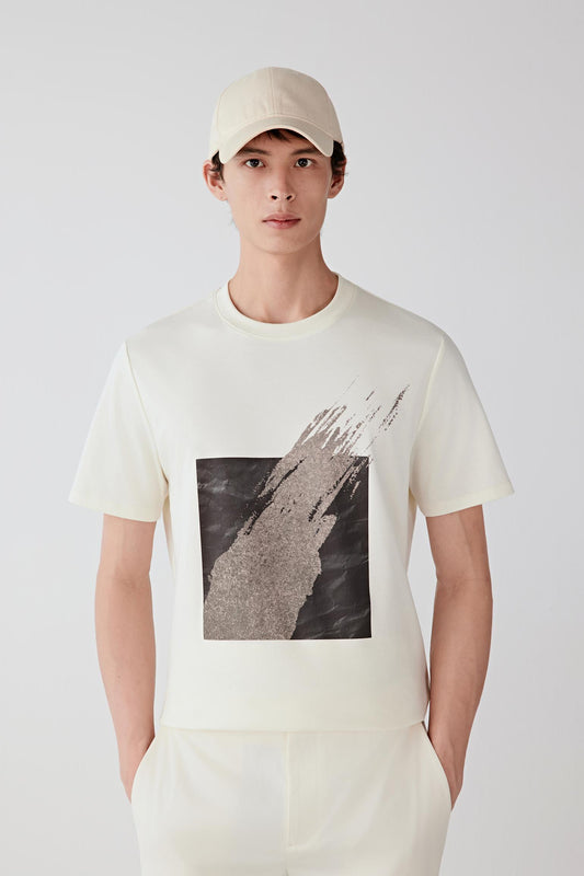 Three-quarter view showing the T-shirt paired with light trousers. The image emphasizes the balanced proportions, short sleeves and structured drape created by the compact interlock knit.
