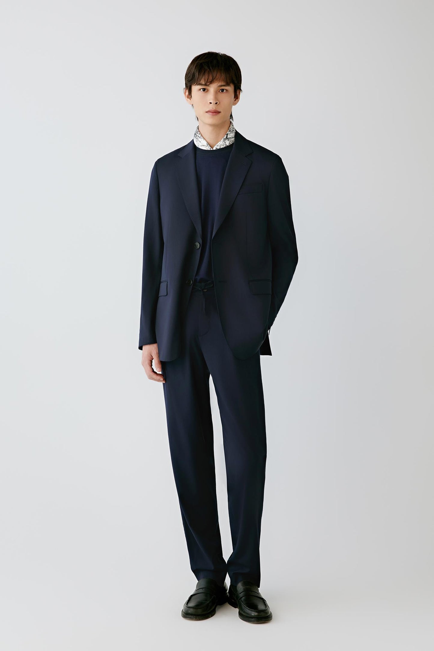 A man modeling a dark navy blue suit with a matching blazer and trousers, worn over a dark crewneck sweater and a white patterned collared shirt, paired with black dress shoes, against a light gray studio background.