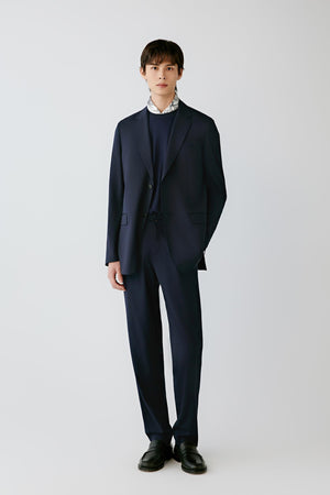 A man modeling a dark navy blue suit with a matching blazer and trousers, worn over a dark crewneck sweater and a white patterned collared shirt, paired with black dress shoes, against a light gray studio background.