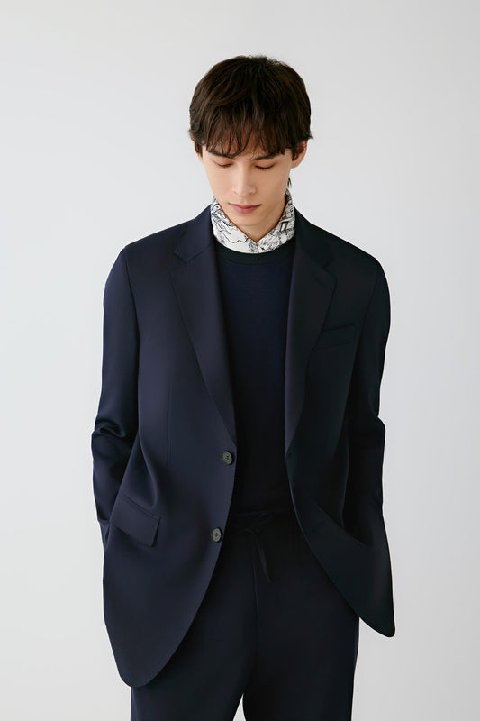 A man wearing a dark navy blue blazer over a matching navy sweater and a patterned white shirt collar, standing against a light gray studio background.