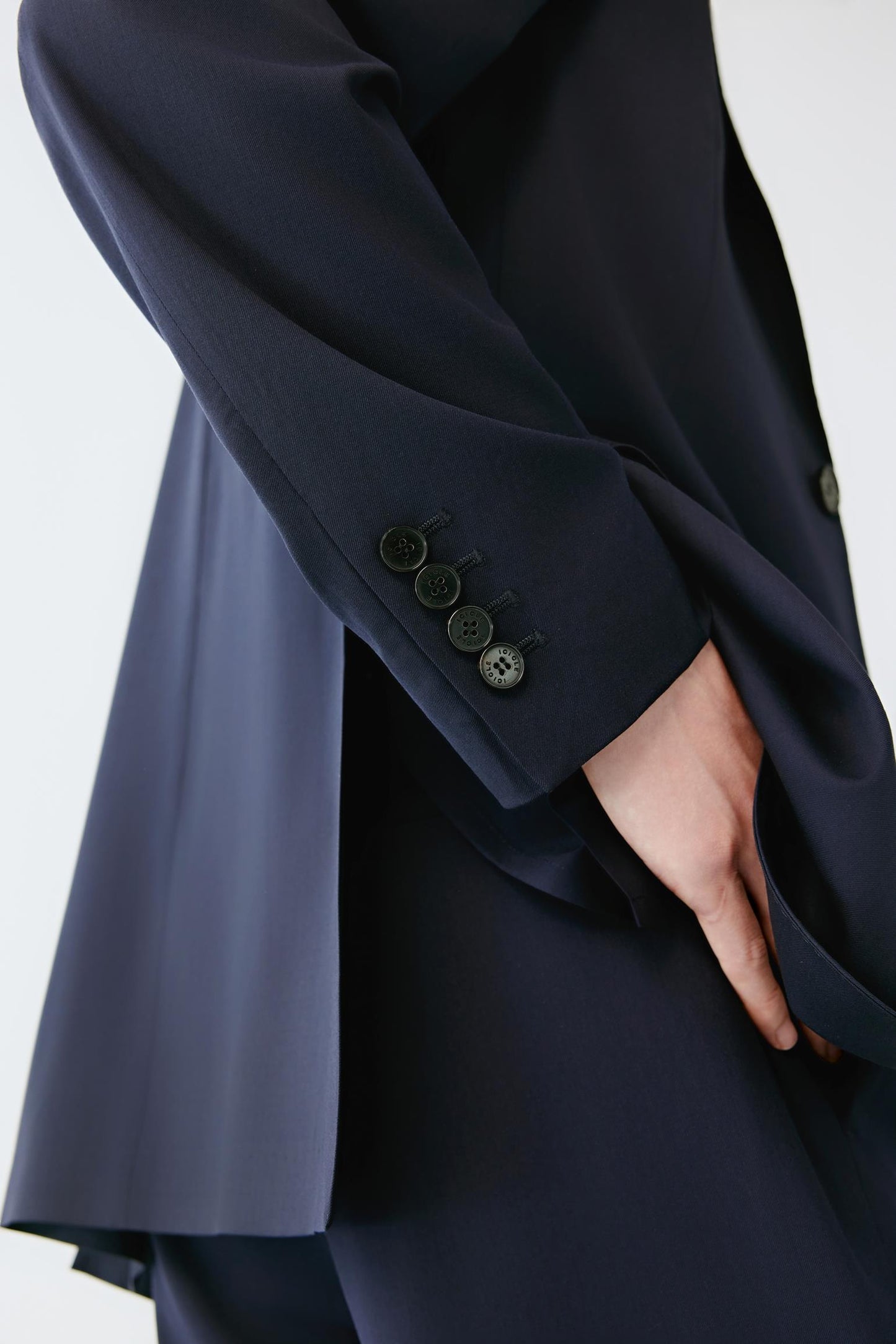 Close-up of a dark navy blue tailored jacket or blazer with a hand resting near the hem, showing the fabric texture and a cuff with four dark buttons against a light gray background.