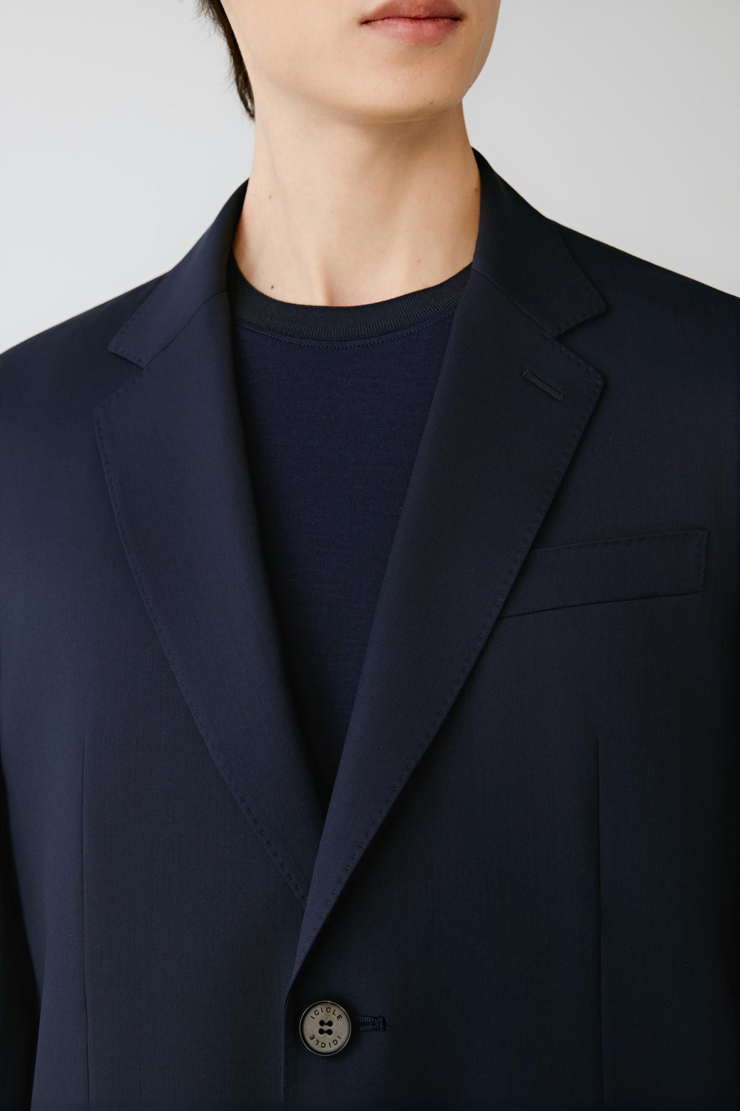 Close-up of a person wearing a dark navy blazer over a matching navy crew-neck shirt against a light gray background.