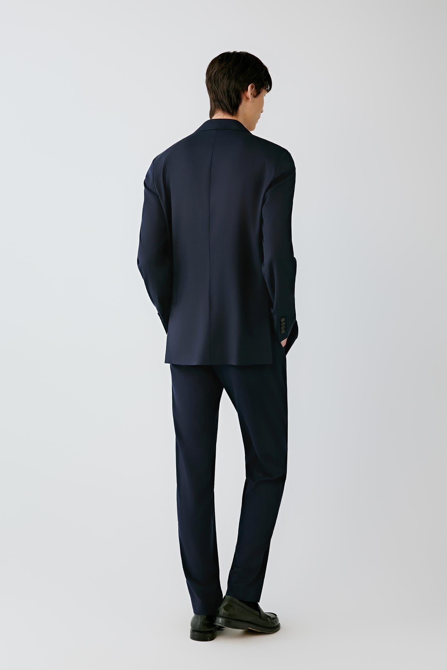 A man wearing a dark navy blue suit, shown from the back, standing against a light gray studio background.