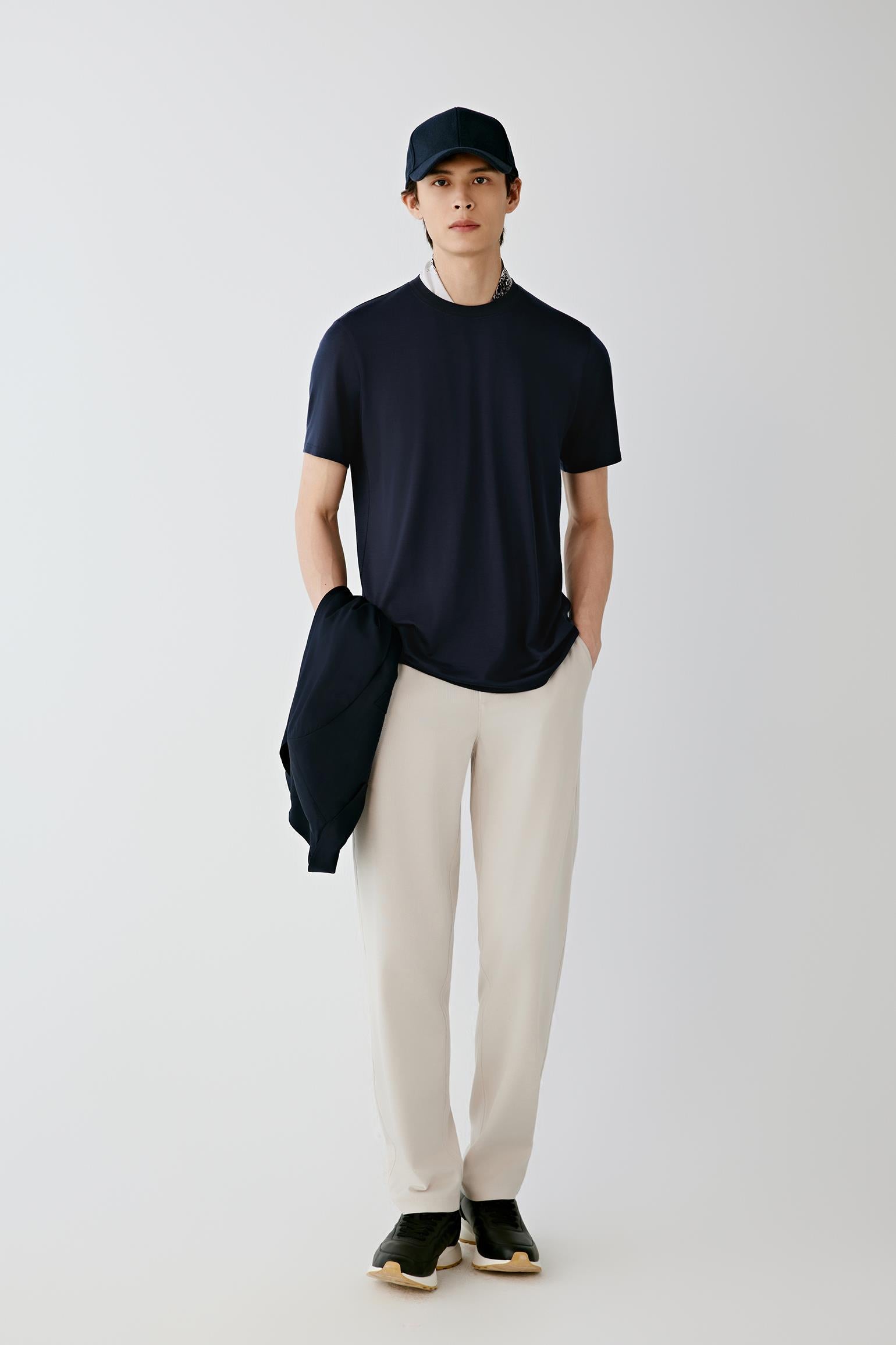 A man stands against a light gray studio background wearing a dark navy short-sleeve crewneck t-shirt, off-white trousers, black sneakers, and a dark navy baseball cap. He is holding a matching navy jacket over his arm.