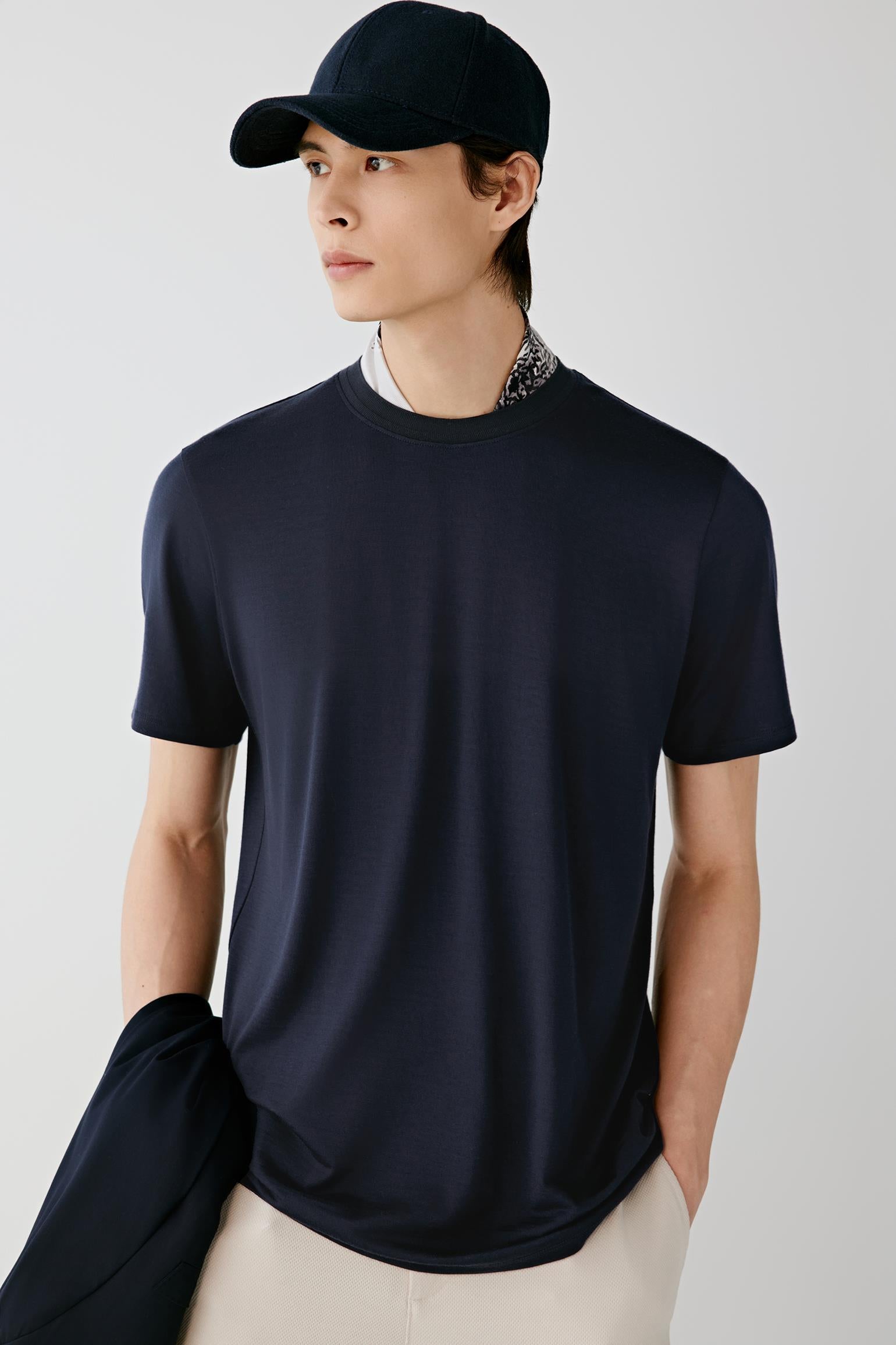 A man wearing a dark navy blue short-sleeve crewneck t-shirt, a matching navy baseball cap, and light beige trousers, standing against a light gray studio background.