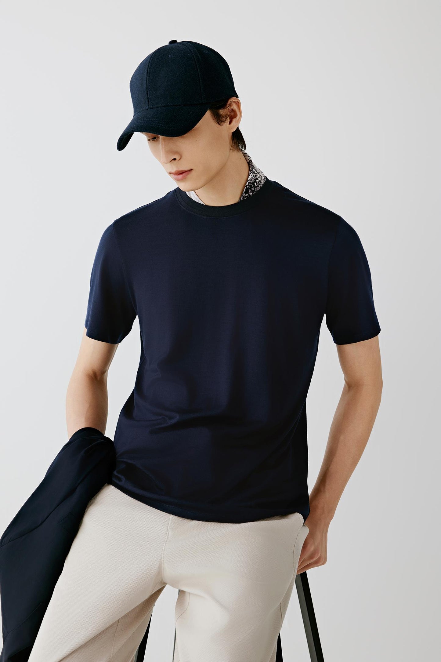 A man wearing a dark navy short-sleeve knit t-shirt, a matching navy baseball cap, and light beige trousers, holding a folded navy jacket against a light gray studio background.