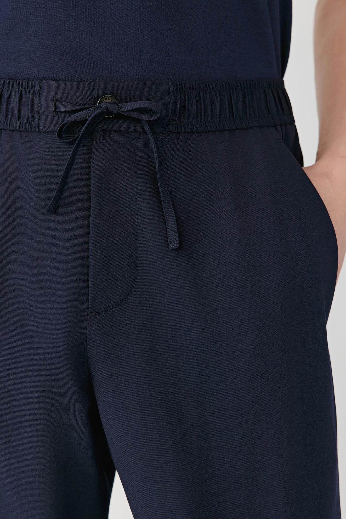Close-up of navy blue drawstring trousers with an elastic waistband and side pocket, worn with a matching navy top against a light gray background.