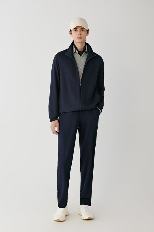 A man modeling a matching navy blue zip-up jacket and trousers set, styled with a light gray collared shirt, a cream baseball cap, and off-white sneakers against a plain light gray studio background.