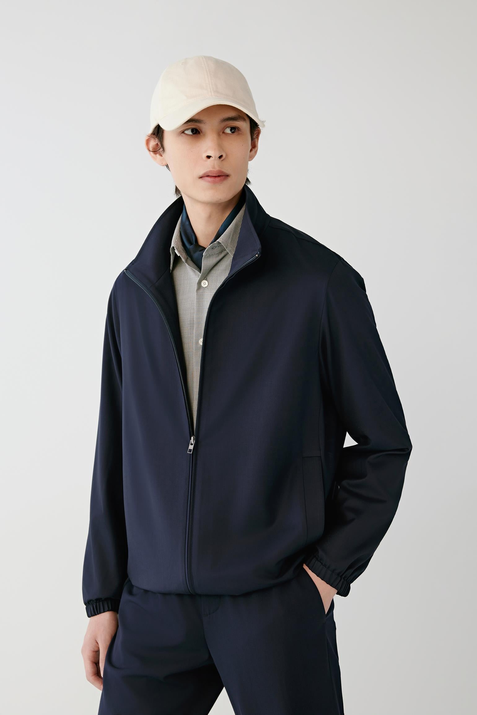 A man wearing a dark navy blue zip-up jacket over a light gray collared shirt, paired with matching navy pants and a beige baseball cap, standing against a light gray studio background.