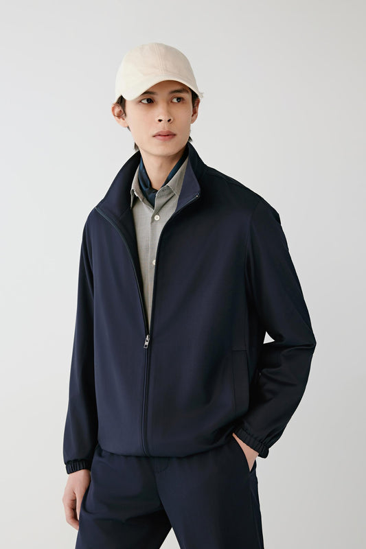 A man wearing a dark navy blue zip-up jacket over a light gray collared shirt, paired with matching navy pants and a beige baseball cap, standing against a light gray studio background.