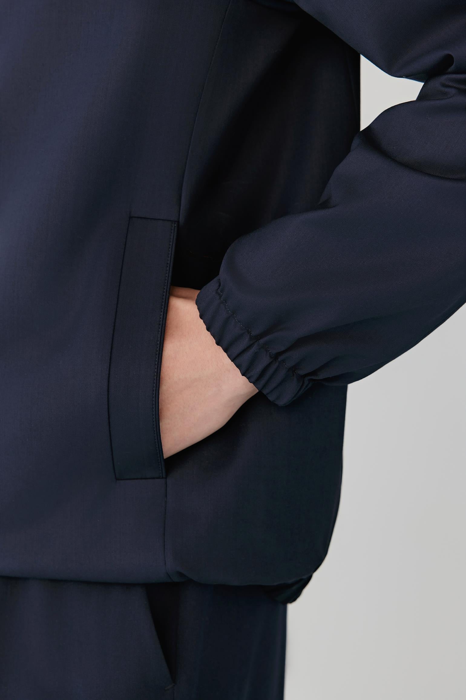 Close-up of a person wearing a dark navy jacket, showing the side pocket with a hand tucked inside and the elasticized cuff of the sleeve, against a light gray background.
