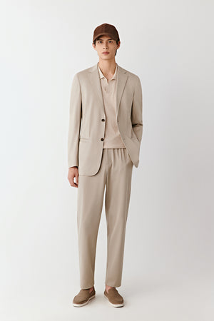 Man wearing a beige piqué cotton tailored jacket with straight cut and two-button closure, styled with matching beige trousers, full-length front view.