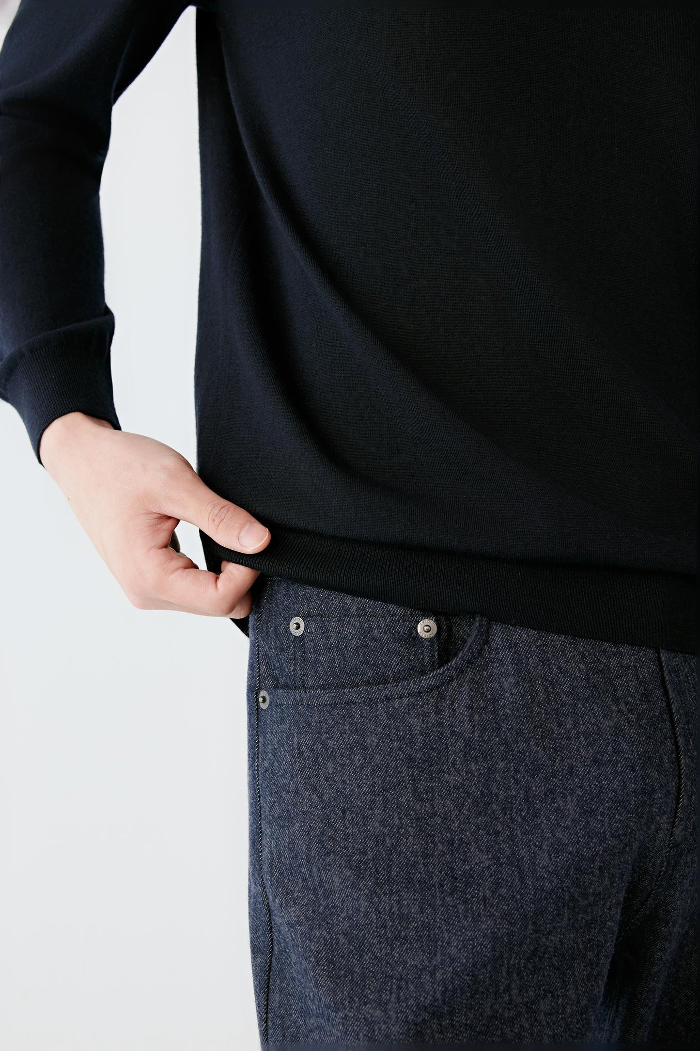 Close-up of a person wearing a black long-sleeve top and dark gray textured trousers, pulling the hem of the shirt slightly with a hand.
