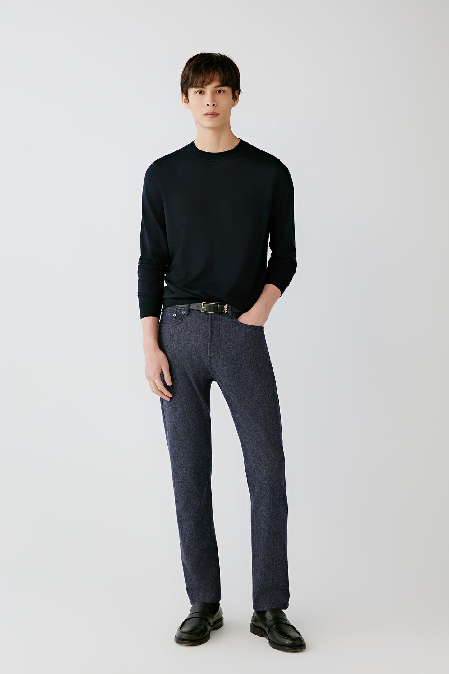 A man modeling a black long-sleeve crewneck sweater and dark gray straight-leg jeans with black leather loafers, standing against a light gray studio background.