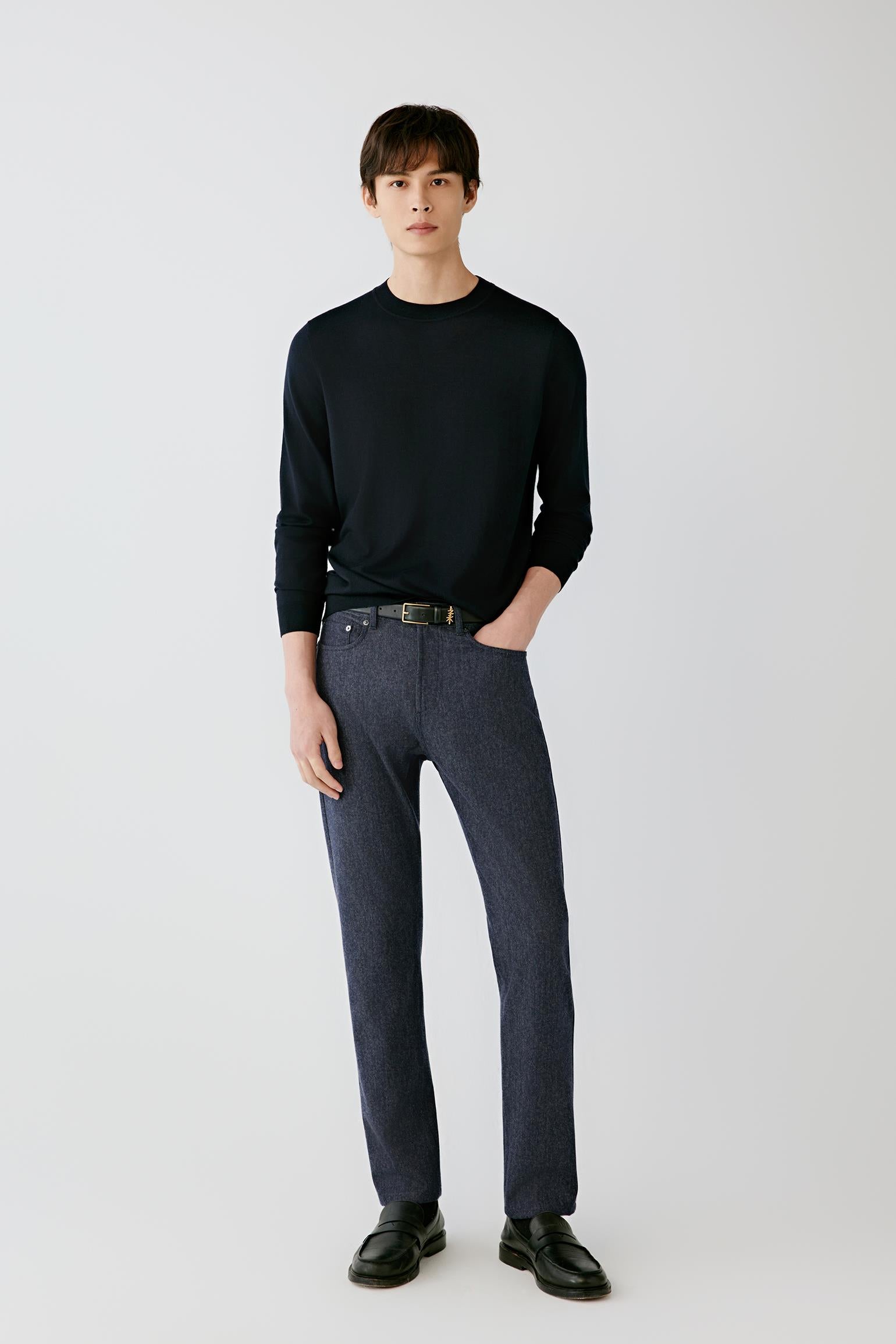 A man modeling a black long-sleeve crewneck sweater and dark gray straight-leg jeans with black leather loafers, standing against a light gray studio background.