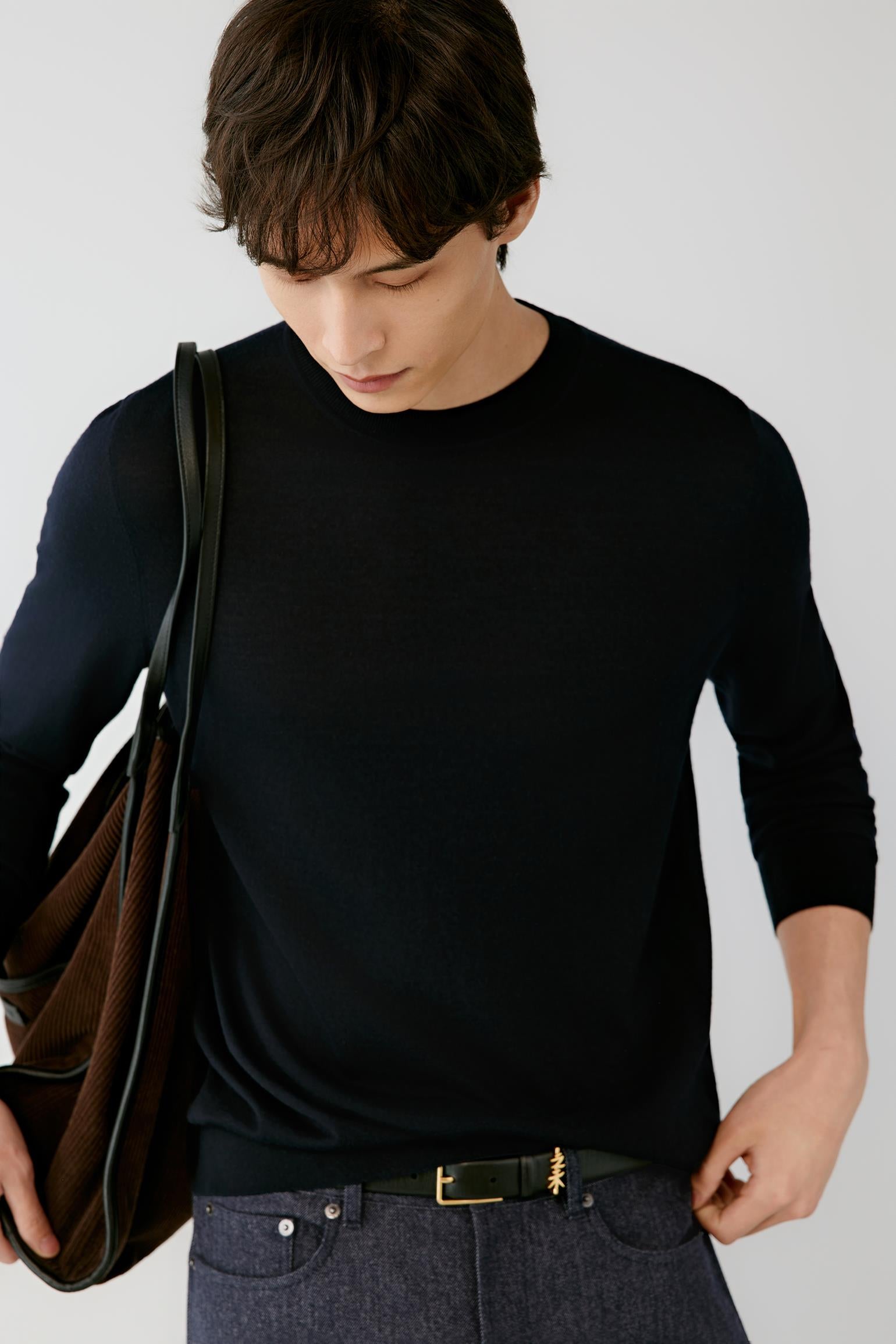 A man wearing a black long-sleeve crewneck sweater, dark gray jeans, and a black belt with a gold buckle, carrying a brown leather shoulder bag against a light gray background.