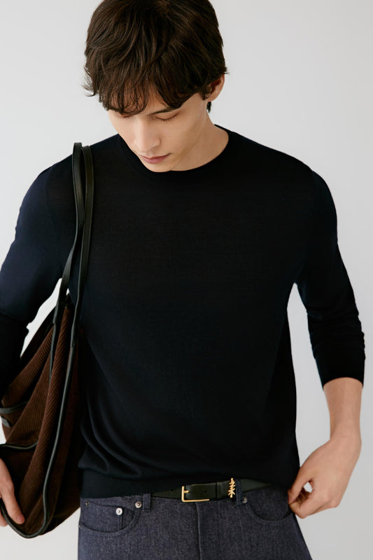 A man wearing a black long-sleeve crewneck sweater, dark gray jeans, and a black belt with a gold buckle, carrying a brown leather shoulder bag against a light gray background.