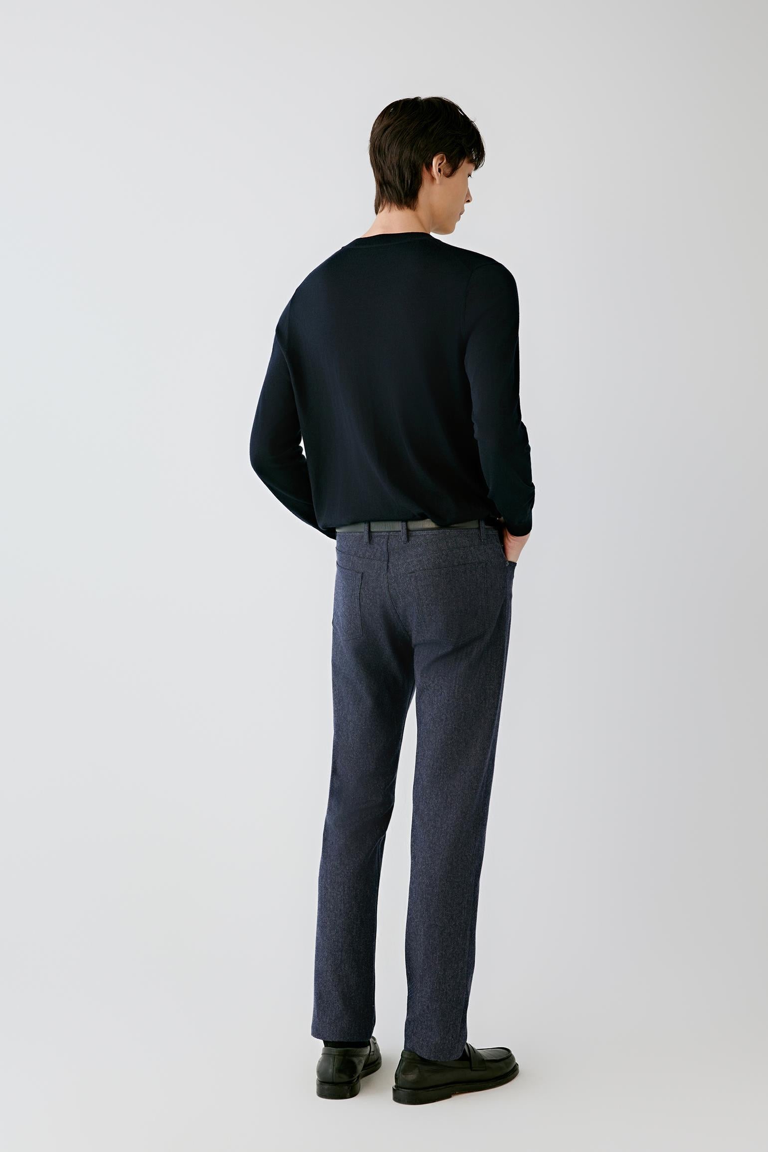A man viewed from behind wearing a dark navy long-sleeve crewneck sweater, dark gray trousers, and black shoes, standing against a light gray studio background.