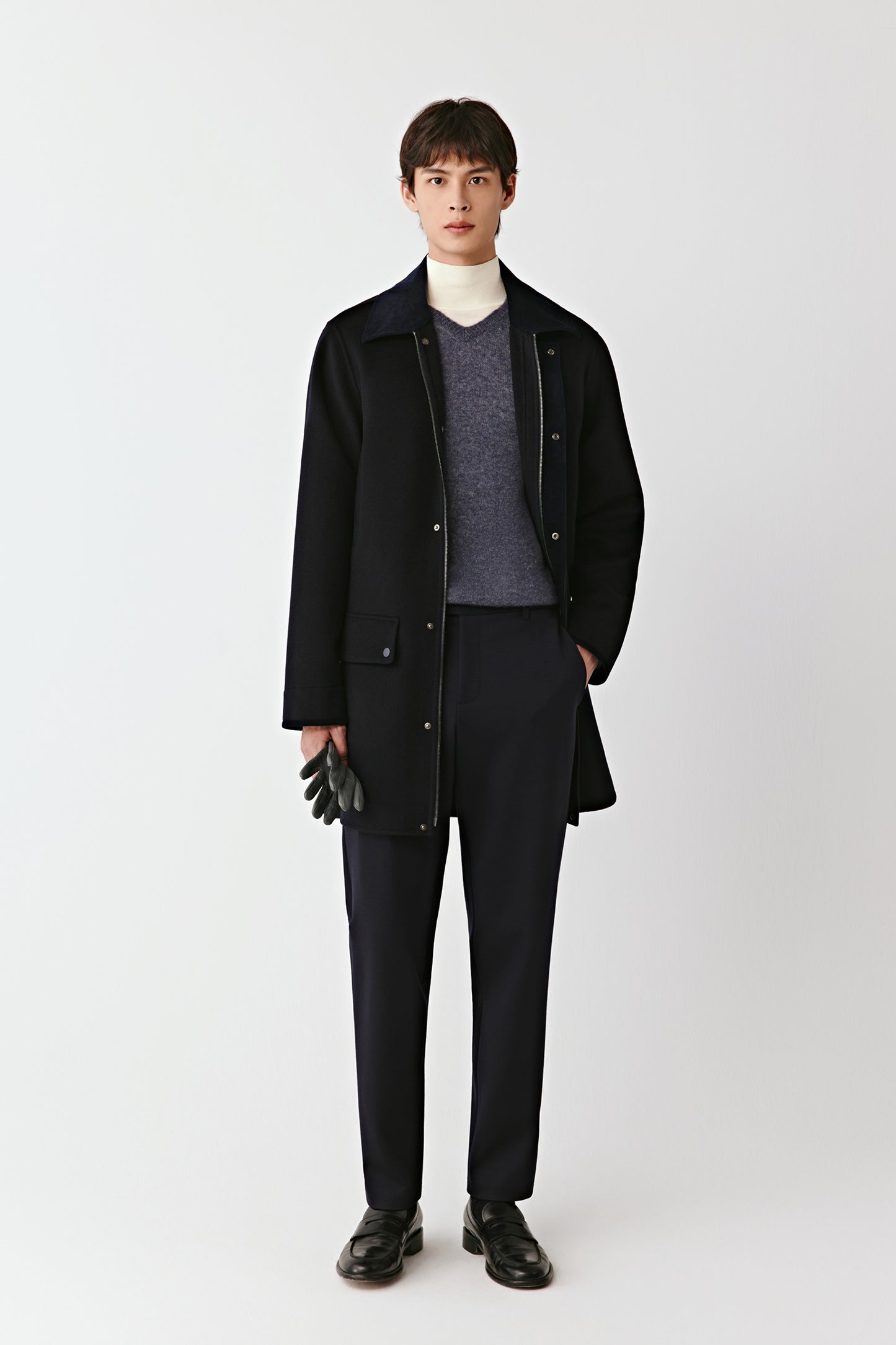 Man wearing a black coat, gray sweater, and black pants on a white background