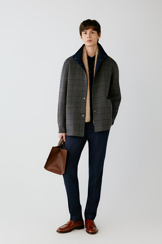 A man modeling a dark gray and black plaid zip-up jacket with a navy blue collar, worn over a beige sweater and dark trousers, holding a brown leather bag against a light gray studio background.