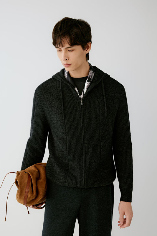 A man wearing a dark charcoal gray ribbed zip-up hoodie with a patterned inner collar, paired with dark trousers and holding a brown suede backpack, against a light gray studio background.