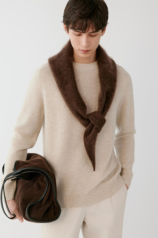A man wearing a beige knit sweater and cream trousers, styled with a dark brown fuzzy neck scarf tied at the chest and holding a dark brown leather bag against a light gray studio background.