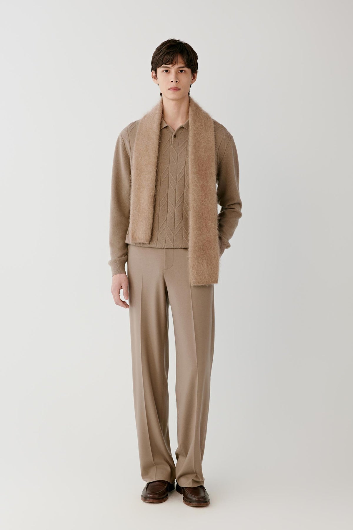A male model stands against a light gray studio background wearing a coordinated beige outfit: a fuzzy sherpa-style collar layered over a knit sweater, paired with matching wide-leg trousers and dark brown loafers.
