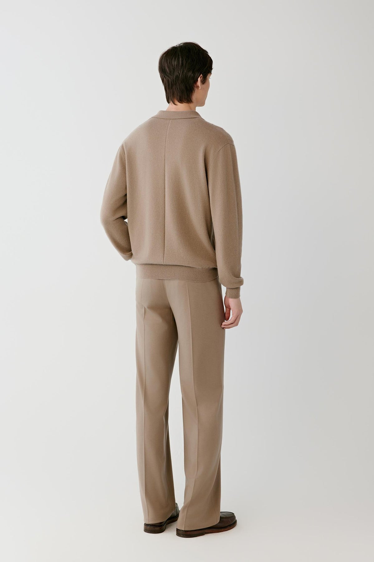A man viewed from behind wearing a matching beige knit sweater and trousers set, standing against a plain light gray studio background.