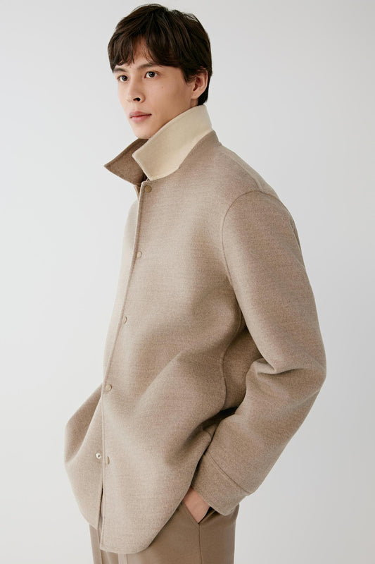 A man wearing a light beige knit cardigan with a high stand-up collar, photographed against a plain light gray studio background.
