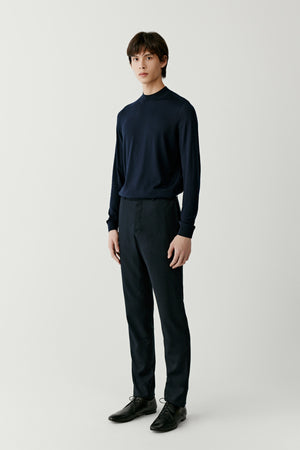 A man stands wearing a navy fine-knit jumper in wool, silk and cashmere with dark trousers, softly lit against a neutral background.