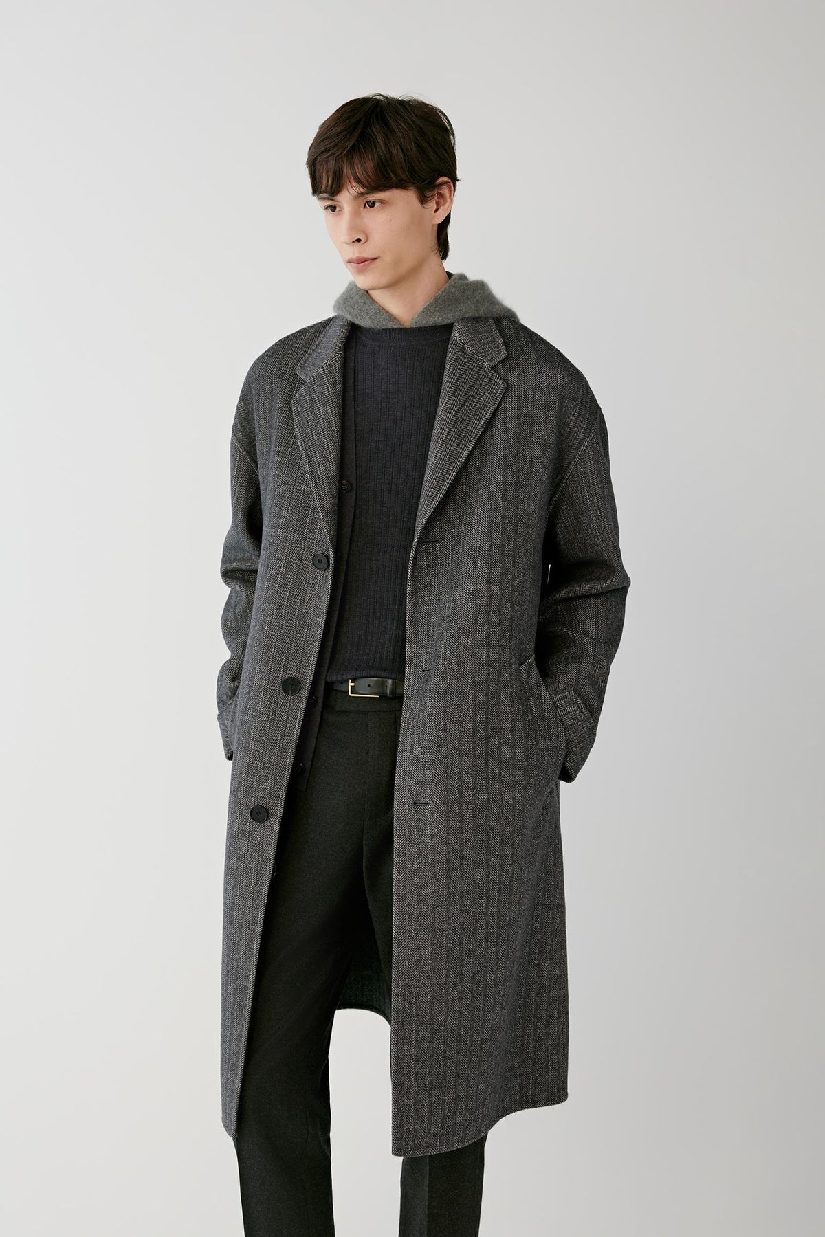 Front view of a man wearing the grey herringbone wool and cashmere coat, highlighting the notched lapels, front fastening and side welt pockets.