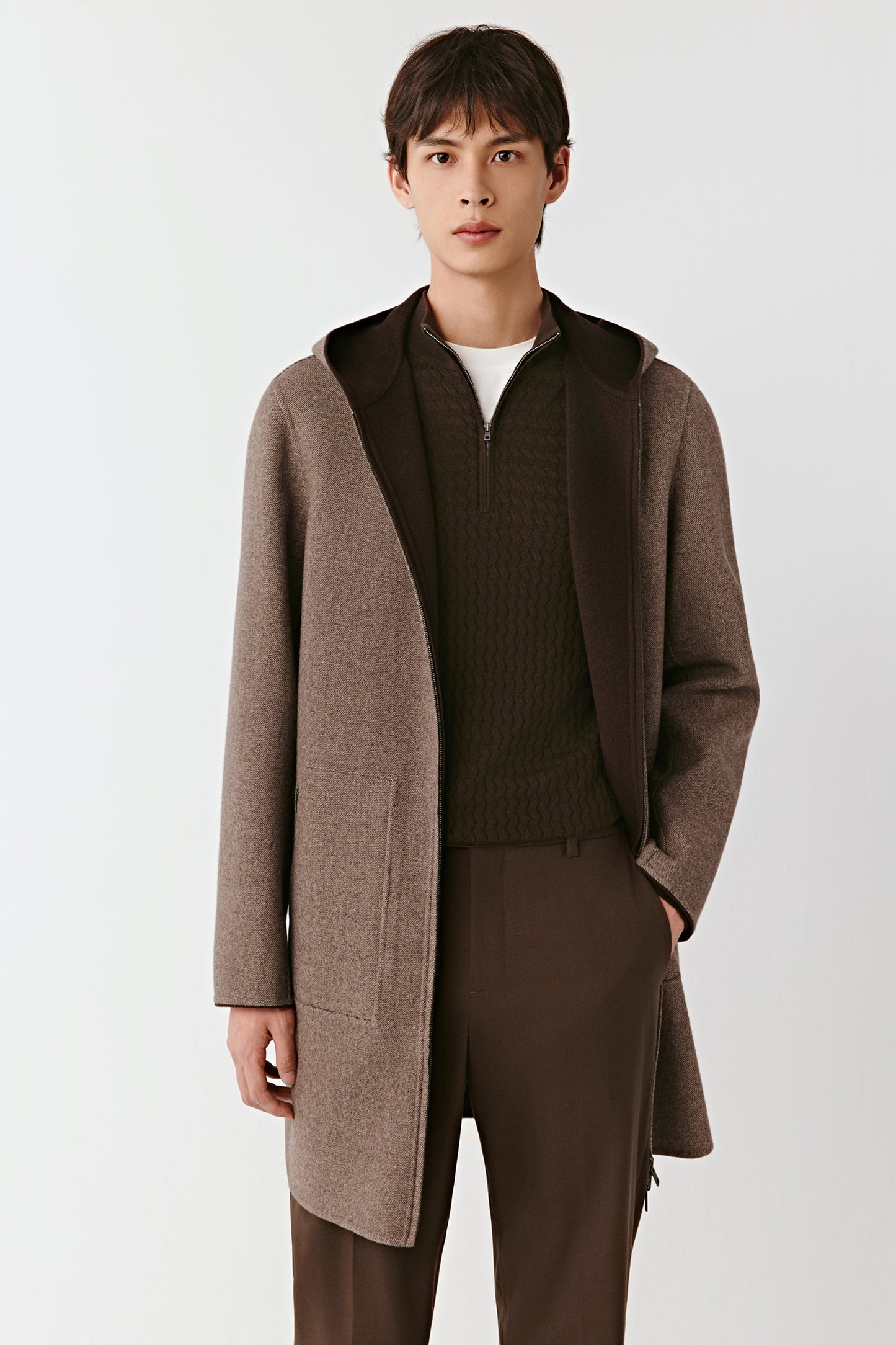 Man wearing a brown coat over a sweater and pants on a white background