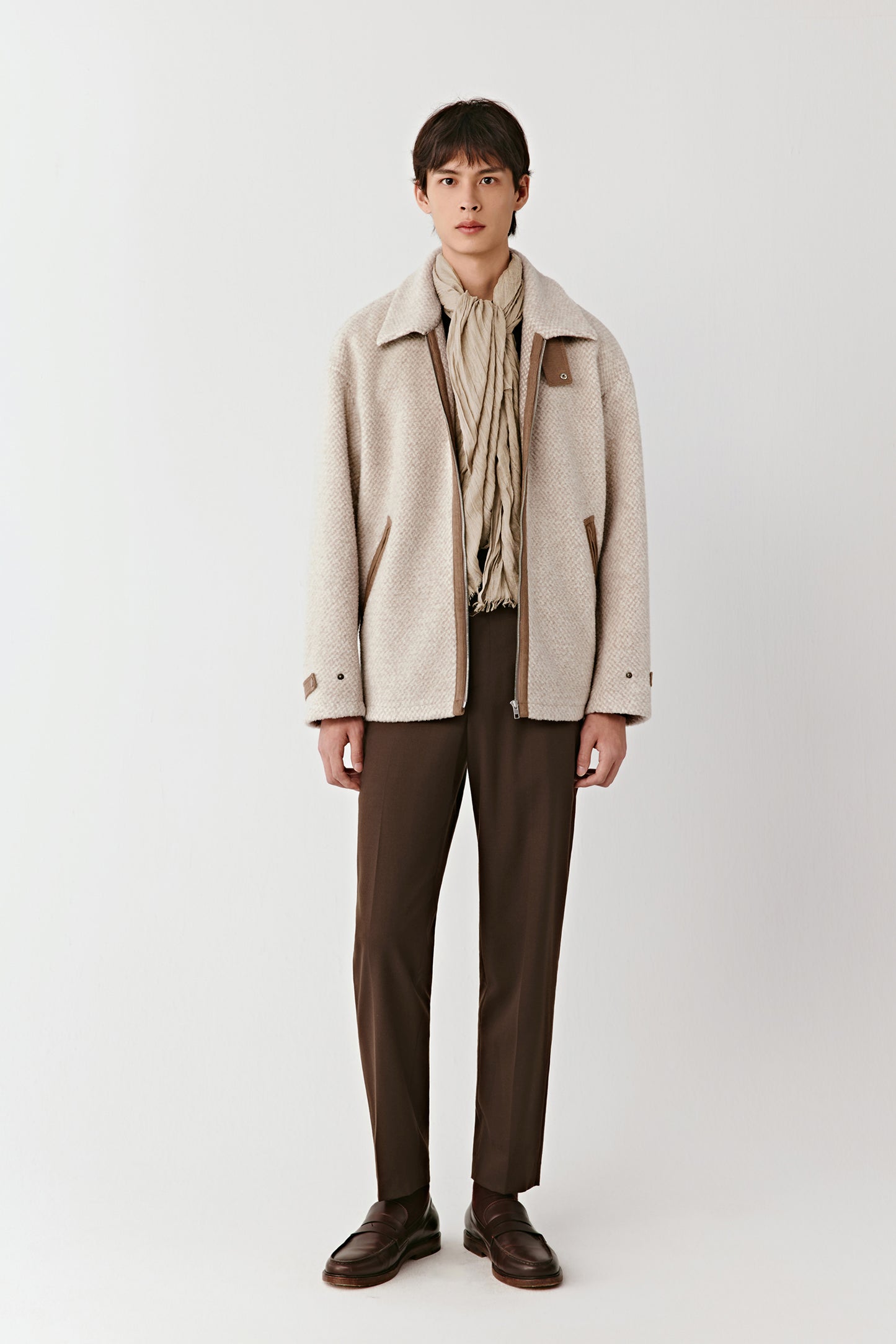 Person wearing a beige coat, beige scarf, and brown pants on a white background
