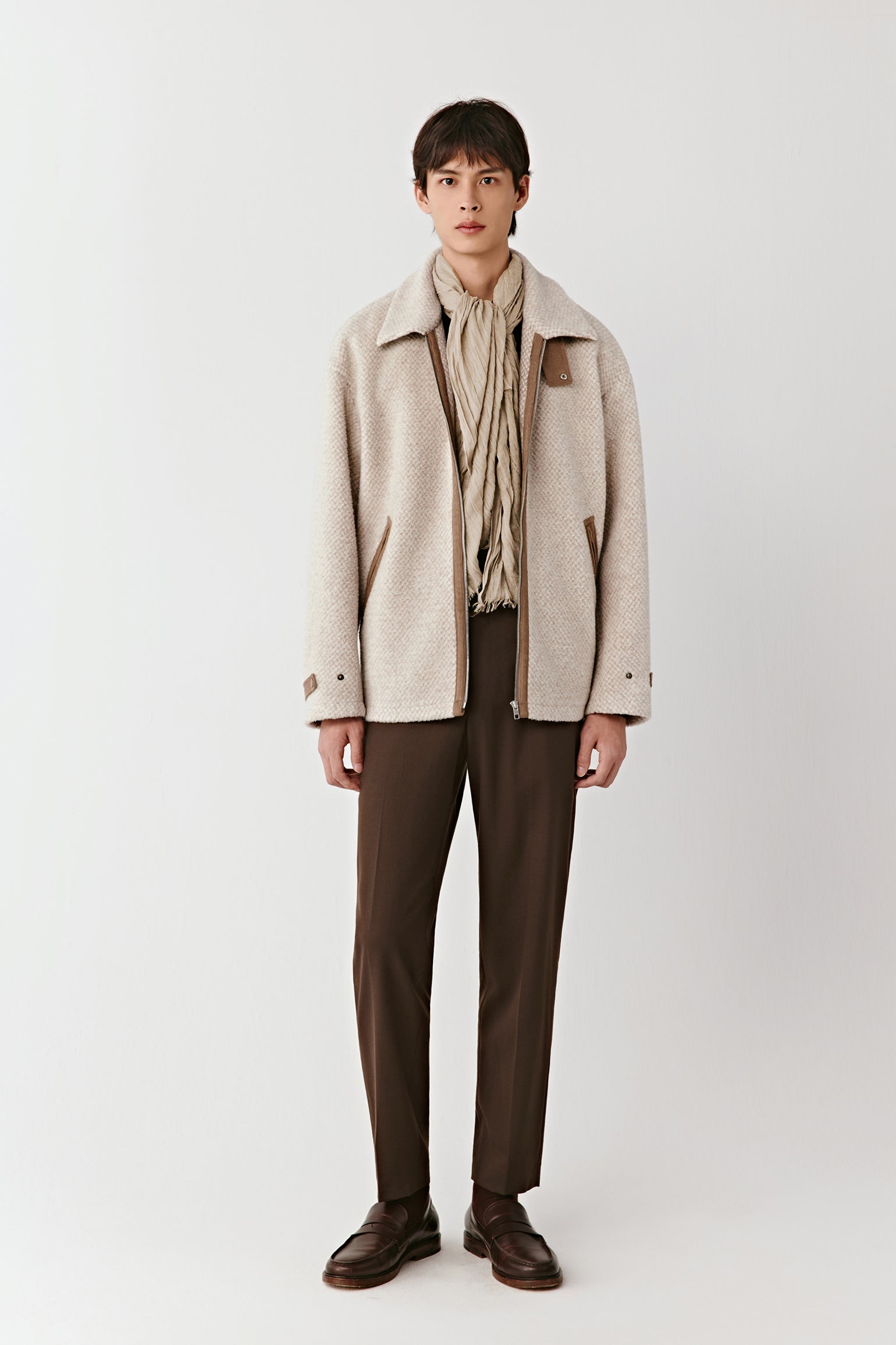 Person wearing a beige coat, beige scarf, and brown pants on a white background