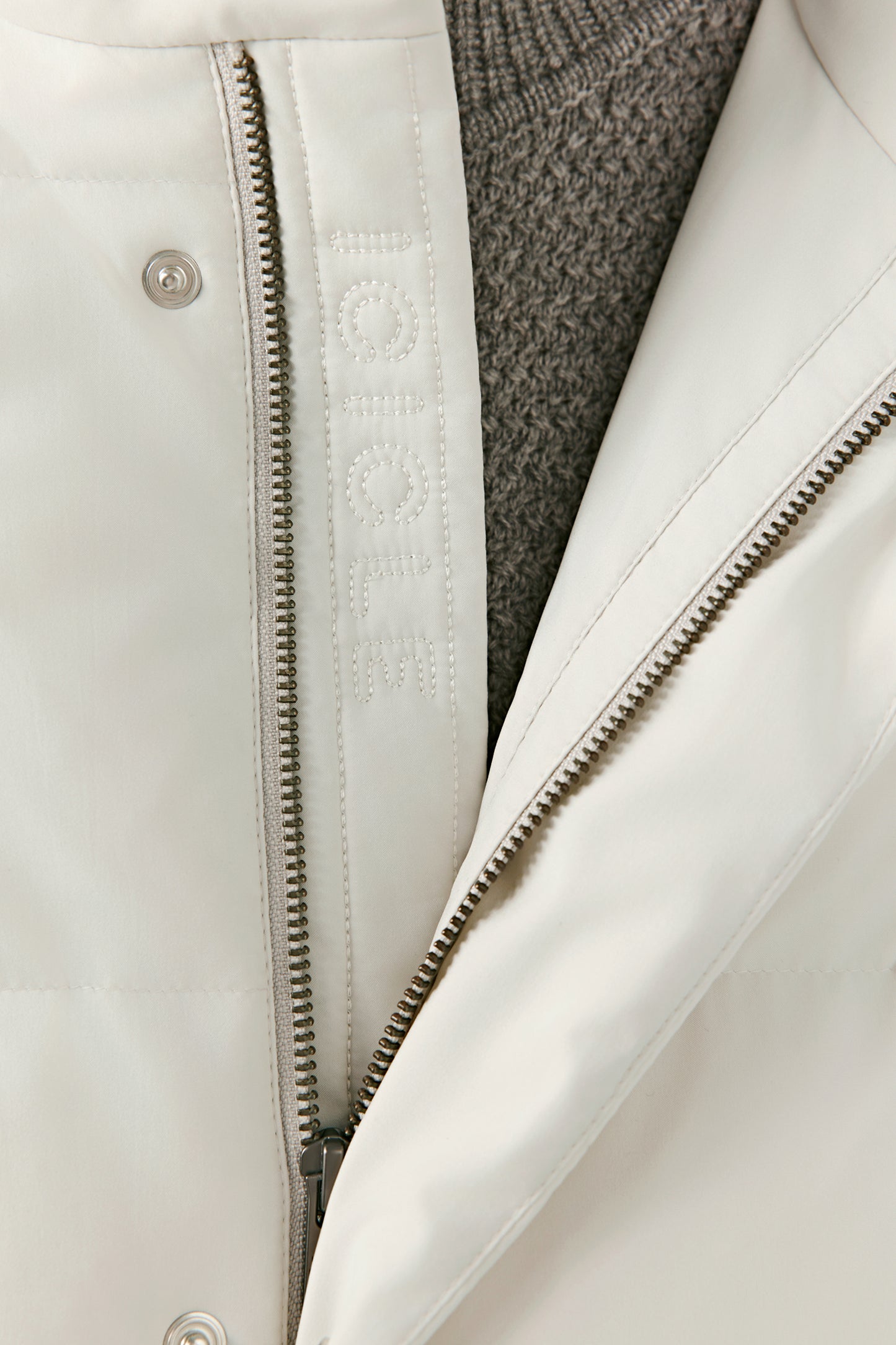 Close-up detail of a cream recycled polyester puffer coat showing the metal zip fastening, snap button, and inner placket with tonal embroidered ICICLE logo, revealing the quilted construction and inner knit layer.
